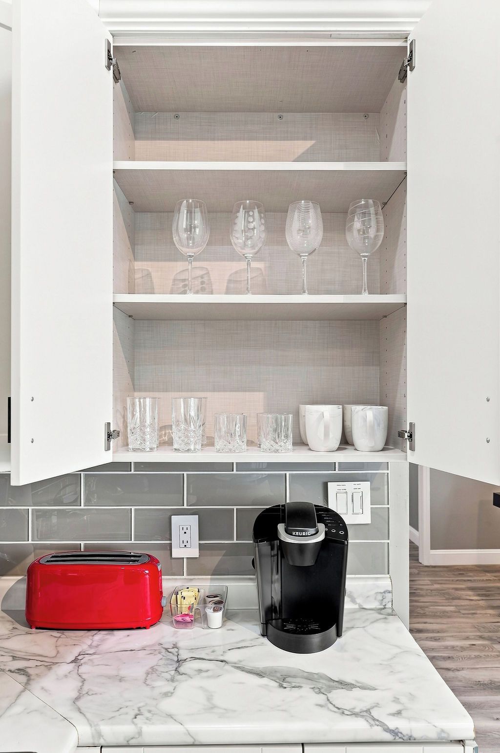 A kitchen with a coffee maker, a toaster, and a cabinet with wine glasses and cups.