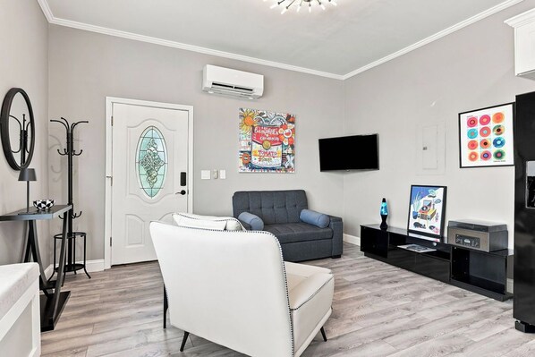 A living room with a grey sofa, a white armchair, a flat-screen TV, and a wall-mounted air conditioner.