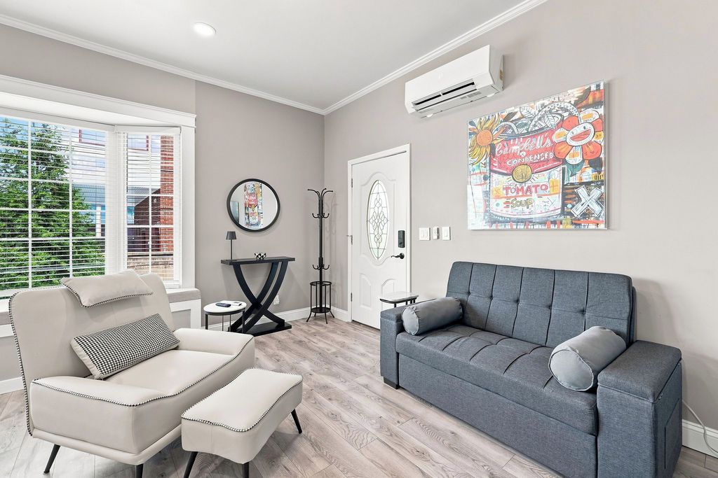 Una sala de estar con un sofá gris, un sillón blanco y una mesita. Hay una ventana con persianas, una puerta y un aire acondicionado de pared.