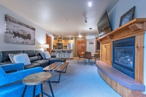 A modern living room with a sofa, coffee table, and chairs, a kitchen in the background, and a fireplace with a TV above it. - Beautiful Teton Creek Resort Condo below Grand Targhee Resort and in Driggs ID (Driggs)