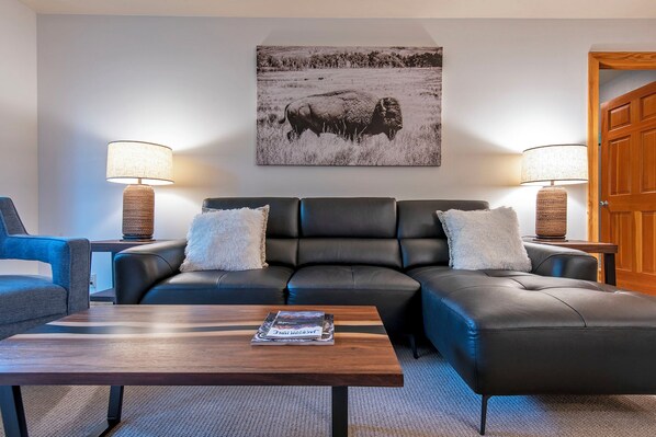 A living room with a black leather sectional sofa, two side tables with lamps, a coffee table, and a framed picture on the wall.