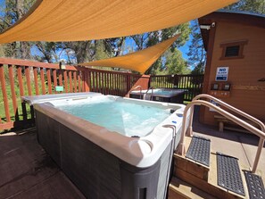A hot tub with a wooden deck and a shaded area, a wooden building with a 'NO GLASS IN POOL AREA' sign, and a wooden fence.