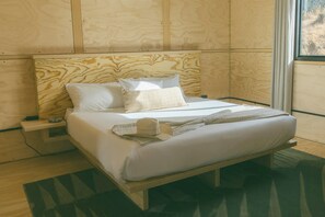 A single bed with a wooden headboard and a nightstand, a window with a view, and a green patterned rug on the floor. - Cobalt Cabin by Homestead Modern (Yucca Valley)