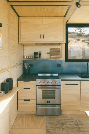 A compact kitchen with wooden cabinets, a stainless steel stove, and a window overlooking a natural landscape. - Cobalt Cabin by Homestead Modern (Yucca Valley)