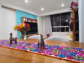 A room with a wooden dining table, colorful patterned rug, decorative animal figurines, a vase with flowers, and a television on a stand. - Moderno Depa Familiar de 3BR con Garaje Cerca a San Blas (Cusco)