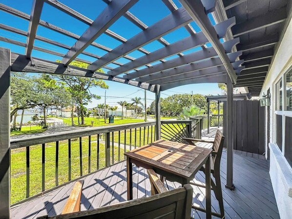 A covered outdoor deck with a table and chairs, overlooking a grassy area with trees and a fence.
