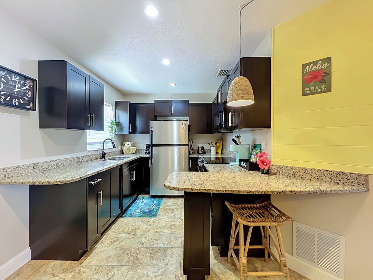 A modern kitchen with dark cabinetry, granite countertops, a stainless steel refrigerator, and a bar area with a wicker stool.