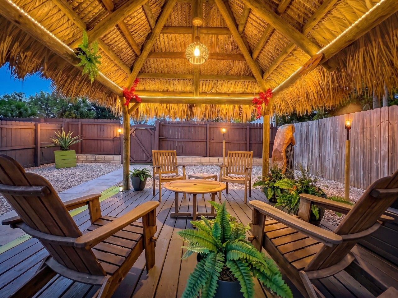 A wooden deck with a thatched roof, surrounded by a wooden fence, and furnished with chairs and a table.