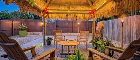 A wooden deck with a thatched roof, surrounded by a wooden fence, and furnished with chairs and a table.