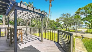 Una terraza de madera con una pérgola, mobiliario exterior y vistas a un parque con palmeras.