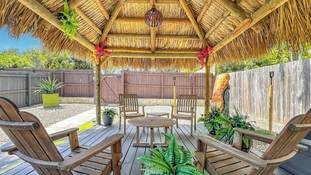 Una terraza de madera con techo de paja, rodeada por una valla de madera, y amueblada con sillas y una mesa de madera.