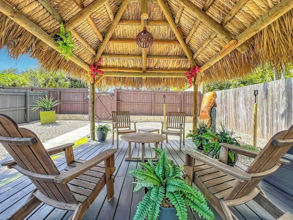 A wooden deck with a thatched roof, surrounded by a wooden fence, and furnished with wooden chairs and a table.