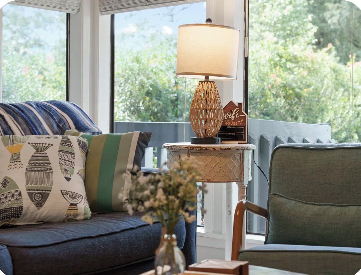 A cozy living area with a sofa, a side table with a lamp, and a view of greenery outside.