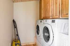 A laundry room with two front-loading washing machines, a yellow vacuum cleaner, wooden cabinets, and a white laundry basket. - The Blue House Hideaway | Less than 20 Minutes to Sugarloaf (Kingfield)