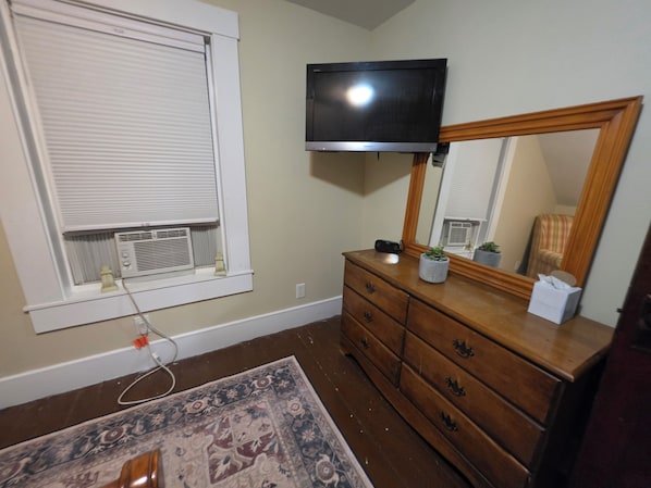 Een kamer met een houten dressoir, een televisie erboven, een raam met jaloezieën en een kamerkleed met patroon op de vloer.