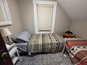 A small, cozy bedroom with a single bed, a nightstand, a lamp, a window with blinds, and a small table with a stuffed toy. - Baxter State Park 2-bedroom house with AC (Millinocket)