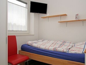 A single-bed room with a wooden frame, a red chair, a television mounted on the wall, and a window with blinds. - Holiday Home in Duhnen With Wi-fi and Parking (Cuxhaven)