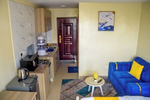 Ein kompakter Wohnbereich mit einer Küchenzeile, einem blauen Sofa mit gelben Kissen und einem Bild an der Wand.