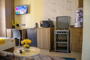 Ein kompaktes Hotelzimmer mit Küchenzeile, einem Bett, einem kleinen Tisch mit einer Vase und einem an der Wand befestigten Fernseher.