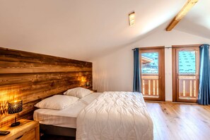 Een slaapkamer met een houten hoofdeinde, een bed met wit beddengoed, een bedlampje en uitzicht op een balkon met blauwe gordijnen.