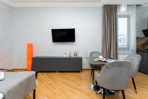 A modern living room with a flat-screen TV mounted on the wall, a grey sofa, a dining table with two chairs, and a large window with curtains. - Zurabyan's Home (Yerevan)