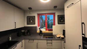 A modern kitchen with white cabinets, black countertops, a red-framed window, and a built-in oven. - Vulkaneifel Artists' House (Mückeln)