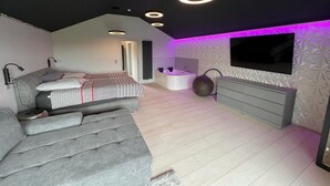A modern bedroom with a grey sofa, a flat-screen TV, a built-in bench, and a ceiling light fixture. - Vulkaneifel Artists' House (Mückeln)
