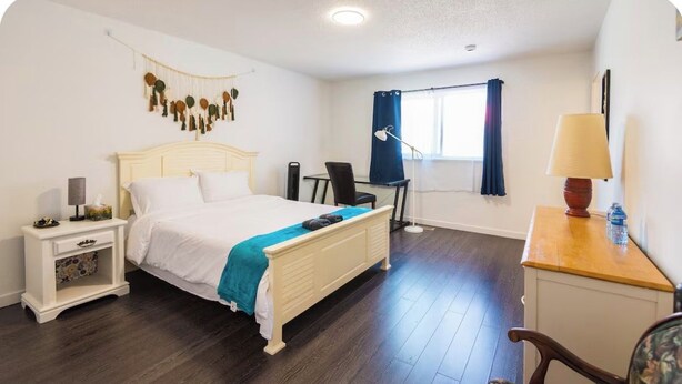 Un dormitorio con cama, mesita de noche, lámpara, escritorio, silla y ventana con cortinas.