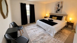 A hotel room with a bed, bedside lamps, a desk with a chair, a window with curtains, and a painting on the wall. - Modern 4-bedroom house  with Country Vibes. (Belton)