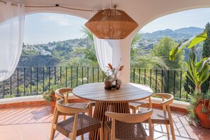 A round wooden table with four chairs, a hanging woven light fixture, and a potted plant on a balcony with a view of a hilly landscape. - Beautiful villa on the Costa Blanca, with a pool, ocean view, garden, and close to the beach and Dénia. (Pego)