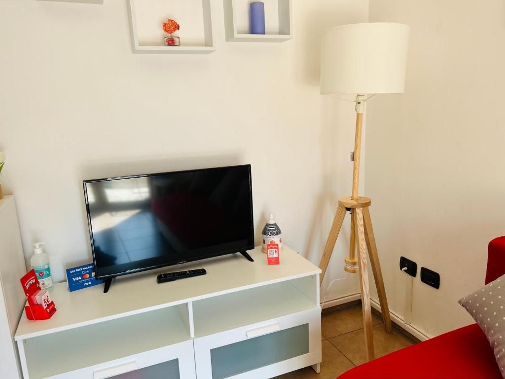 Ein Zimmer mit einem Flachbildfernseher auf einem weißen TV-Schrank, einer Lampe, einem roten Sessel und einem kleinen Regal mit Dekoration.