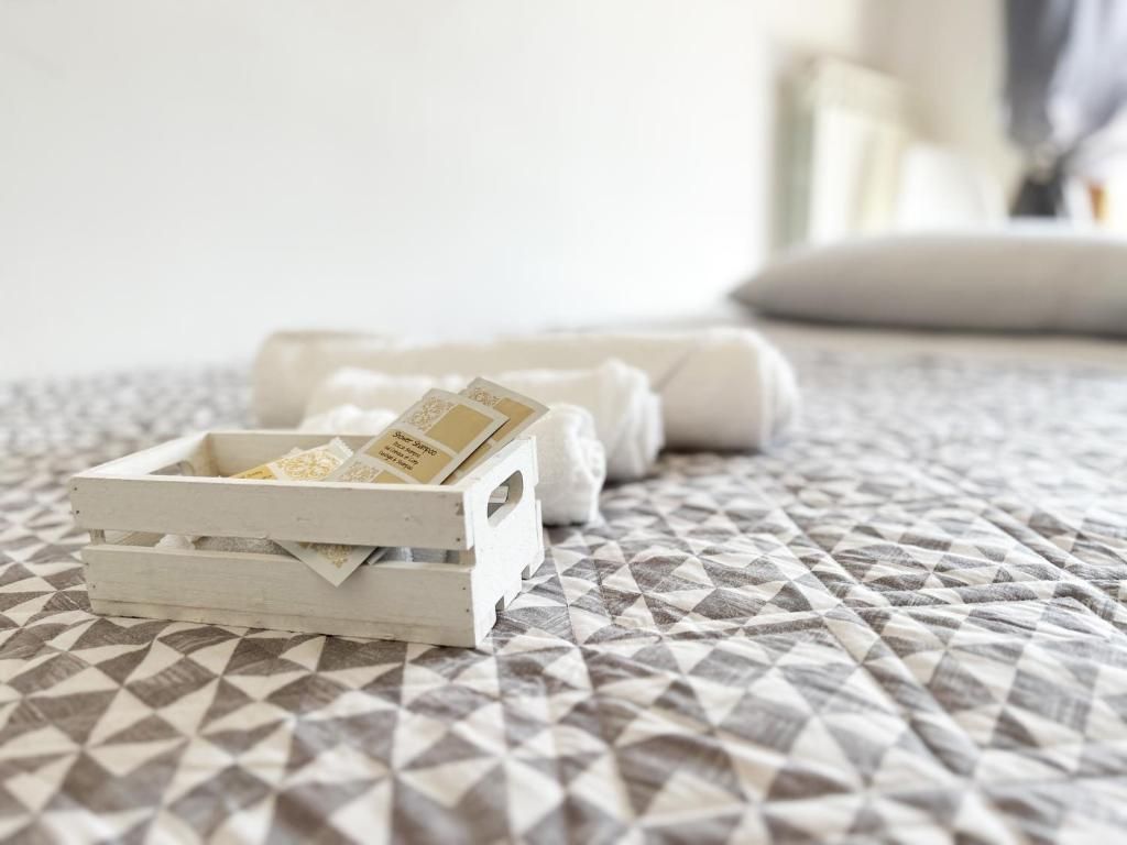 Ein Bett mit einem gemusterten Überwurf, ein weißer Nachttisch mit einer kleinen weißen Schachtel mit Gegenständen und ein aufgerolltes weißes Handtuch.