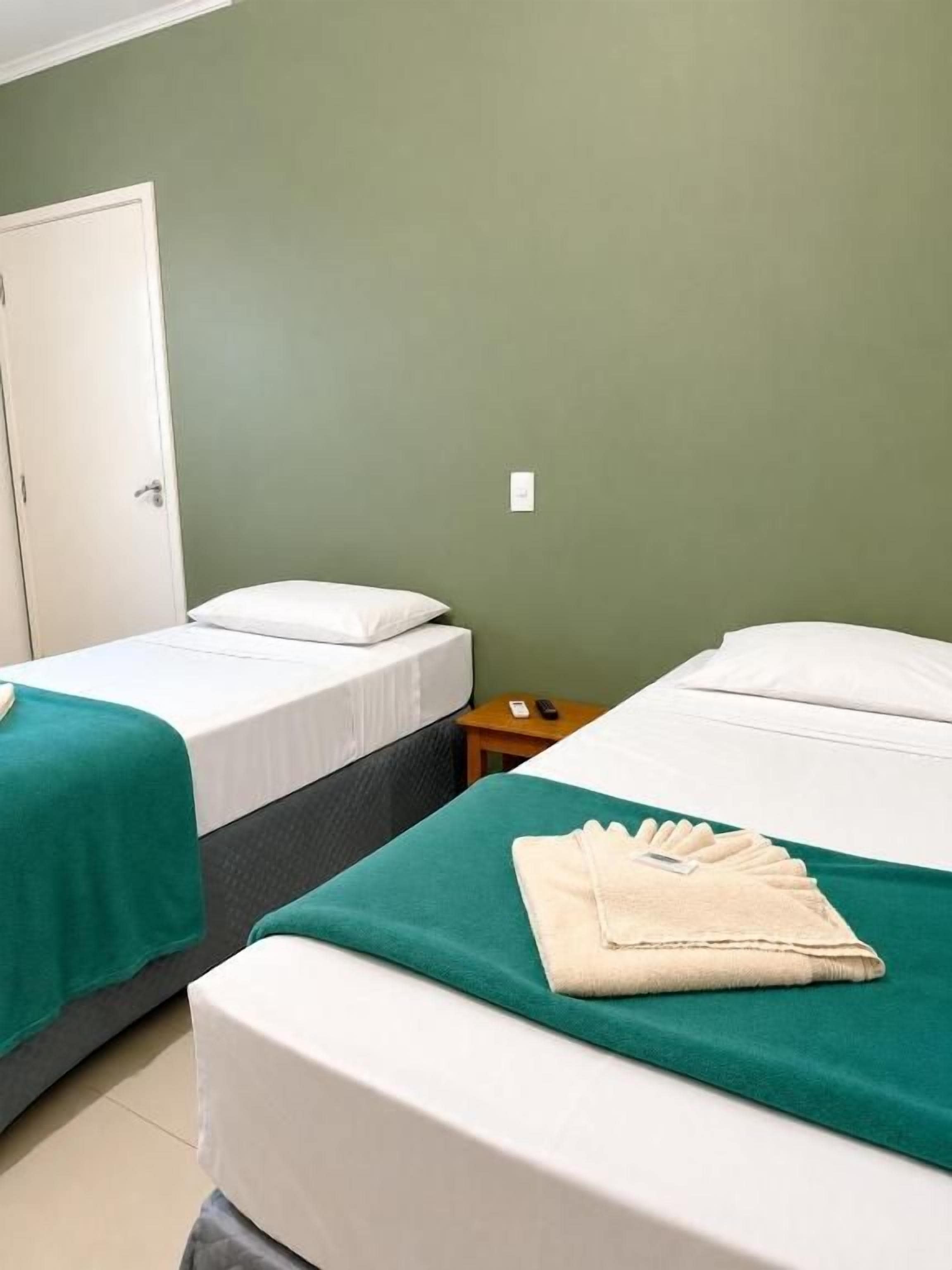 Ein Zimmer mit zwei Betten, jeweils ausgestattet mit einer weißen Matratze und einer grünen Bettdecke, einer weißen Tür und einem kleinen Nachttisch mit Lampe.