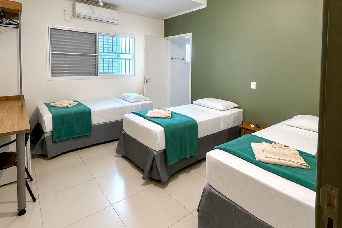A hotel room with three single beds, a window with blinds, a door, and a desk with a chair.
