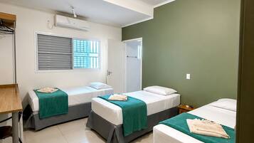 A hotel room with three single beds, a window with blinds, a door, and a desk with a chair.
