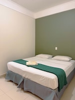 Una cama individual con una manta verde y una toalla doblada, ropa de cama blanca y falda acolchada.