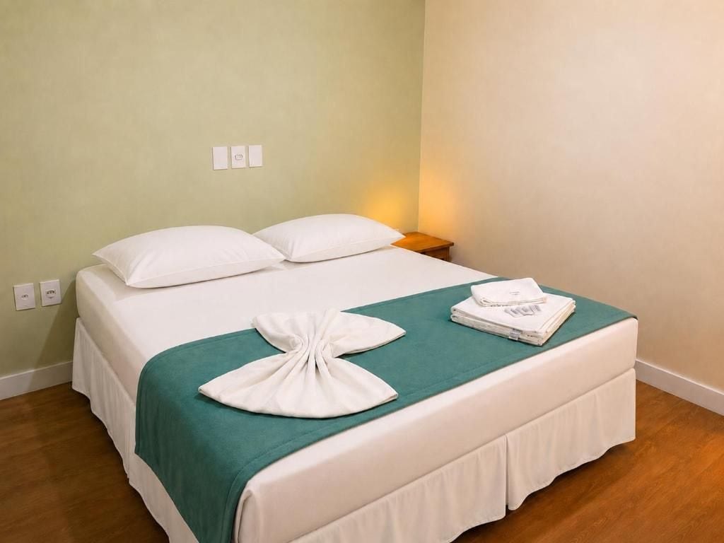 A neatly made bed with white bedding and a teal throw, a folded towel, and a bedside lamp on a wooden nightstand.