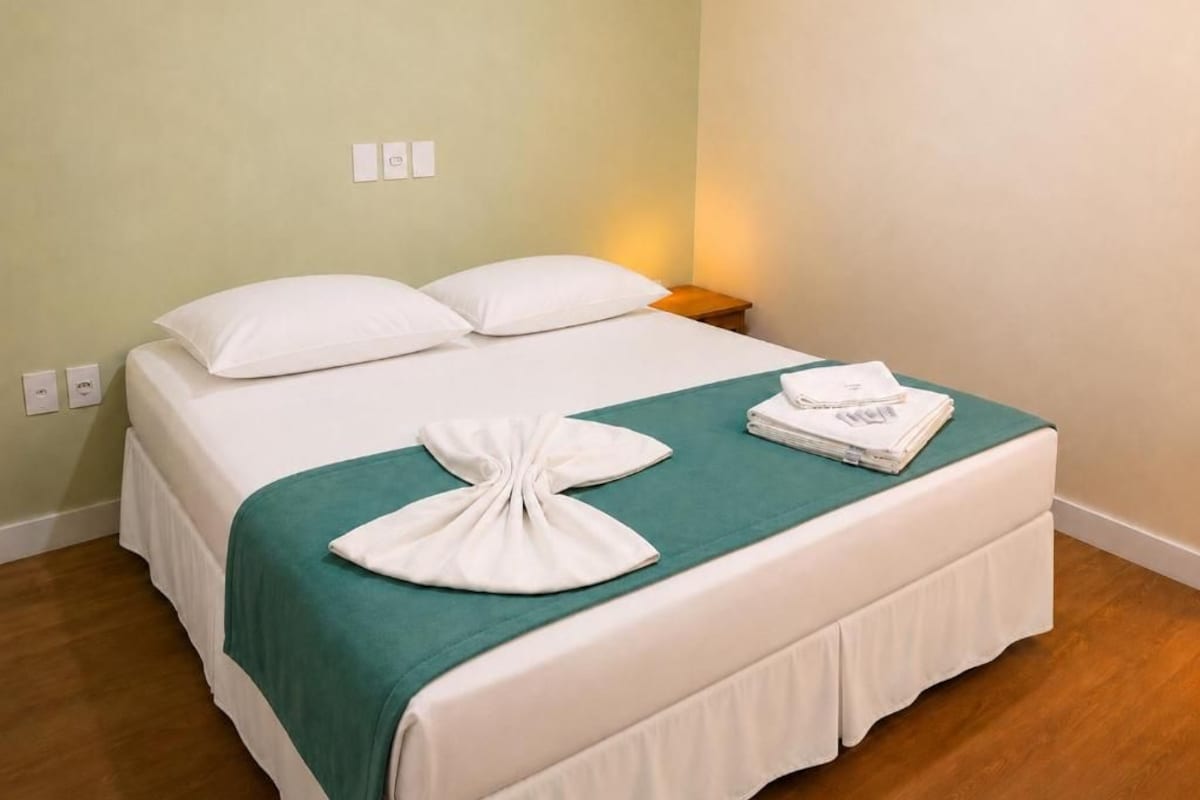 A neatly made bed with white bedding and a teal throw, a folded towel, and a bedside lamp on a wooden nightstand.