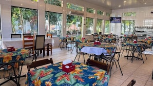 Un comedor interior con mesas cubiertas de estampados tropicales, ventanas amplias y ventiladores de techo.