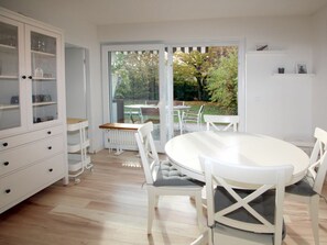 Eine moderne Küche mit weißen Schränken, einem runden Esstisch und Blick auf den Garten durch große Fenster.