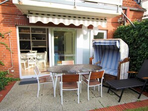 A patio with a wooden table and chairs, a wicker chair, and a glass door leading to an indoor area with a white chair and a bookshelf. - Fasanenweg 2b, Holiday Apartment 2 (Wyk auf Foehr)