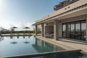 Un espace piscine extérieur moderne, doté de chaises longues et de parasols, d’un patio couvert avec des sièges, et offrant une vue sur le paysage.