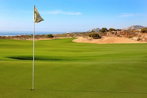 Ein Golfplatz mit Fahne, Fahnenmast und weiter Aussicht auf das Meer und Hügel.