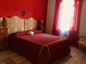 A bedroom with a large bed, a headboard with a decorative pattern, a bedside table with a lamp, and a window with red curtains. - Tramonti d'Agrumi Annex (Naso)