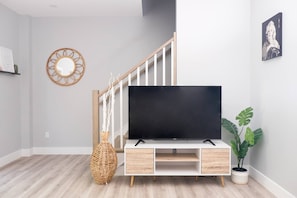 A modern living room with a flat-screen TV on a wooden stand, a staircase, a decorative mirror, and a potted plant.