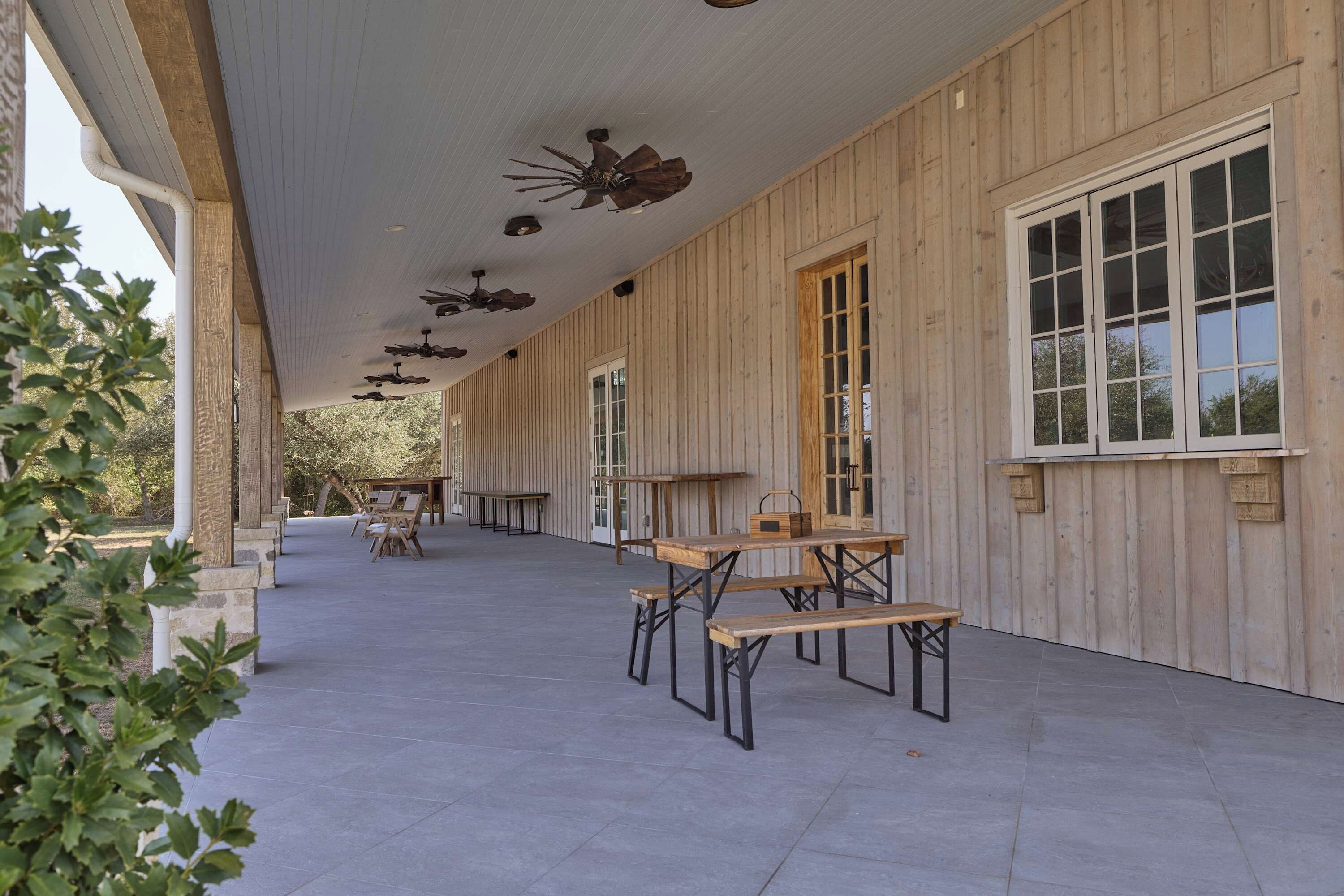 An outdoor covered patio area with wooden tables and chairs, a ceiling fan, and large windows.