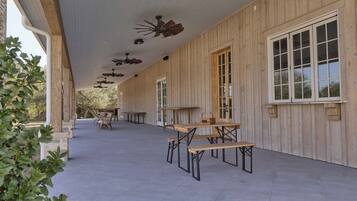An outdoor covered patio area with wooden tables and chairs, a ceiling fan, and large windows.