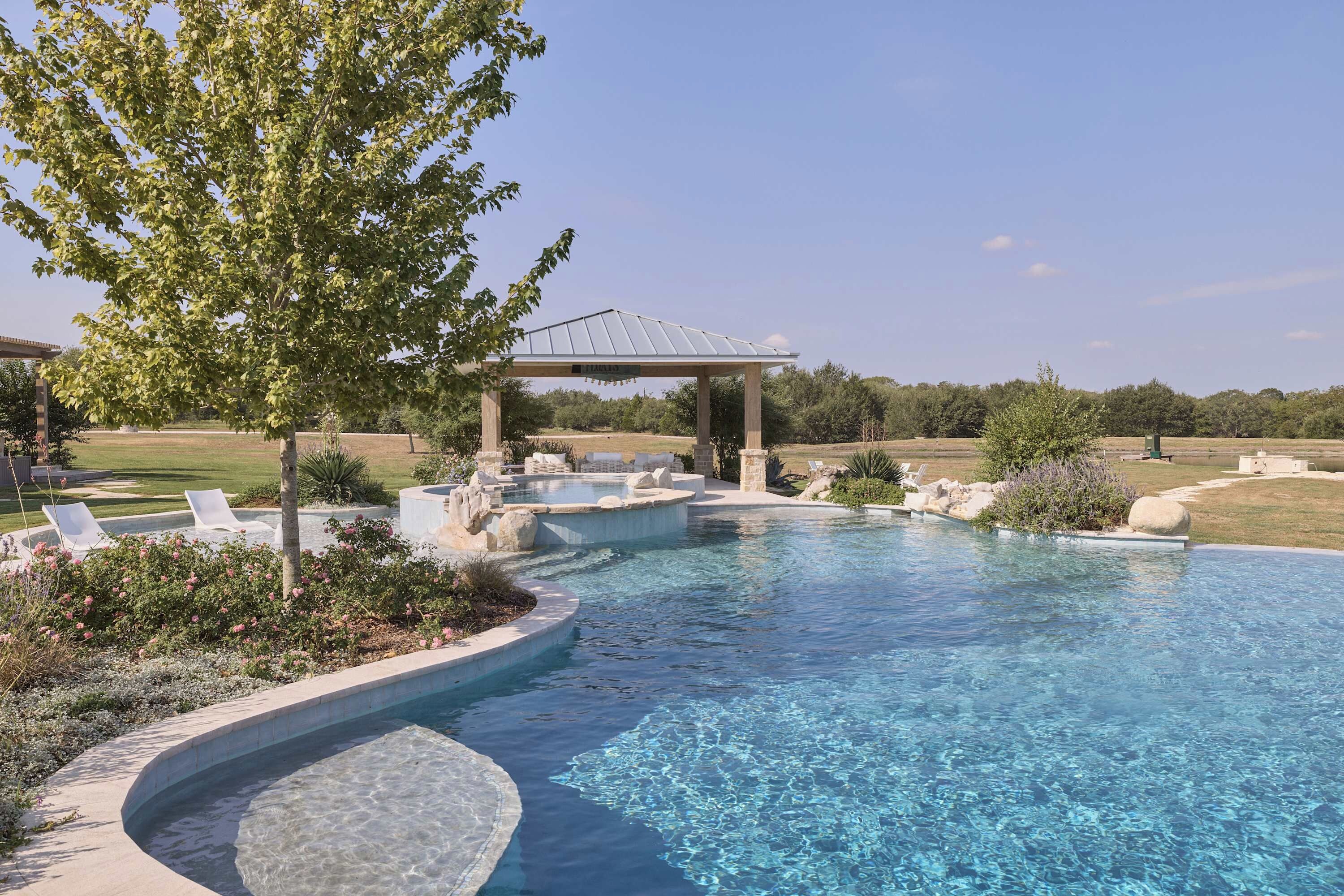A clear swimming pool with a pergola and lounge chairs, surrounded by landscaped gardens and a large tree.