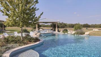 A clear swimming pool with a pergola and lounge chairs, surrounded by landscaped gardens and a large tree.