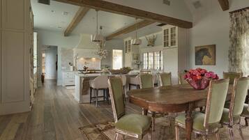 A spacious kitchen with high ceilings and exposed beams, a dining area with a wooden table and vintage chairs, and a large island with a sink and fruit bowl.